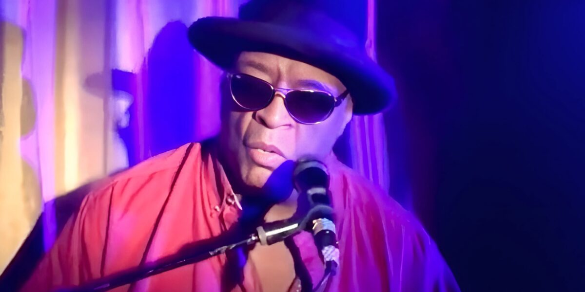 Photo of Ricky Williams. The image shows a musician performing on stage under purple and blue lighting. The musician is wearing a dark hat and sunglasses, which adds a sense of style and mystique to the performance. Dressed in a red shirt, they are focused intently on the microphone and instrument in front of them, capturing a moment of musical expression. The ambiance of the stage is intimate and soulful, reflecting the essence of a live jazz performance.