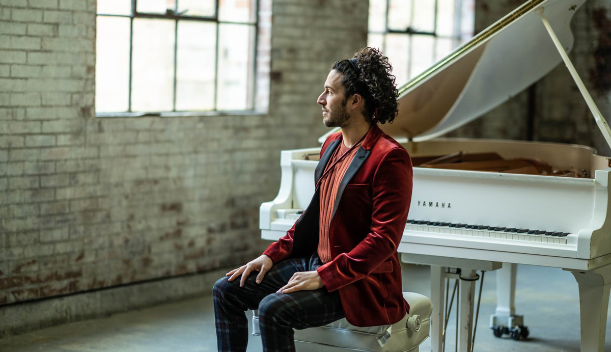 A person named Emmet Cohen is sitting at a piano, showcasing their connection to music. The setting is indoors, likely within a building that features musical elements. The image highlights themes of clothing and the musical instrument being played.
