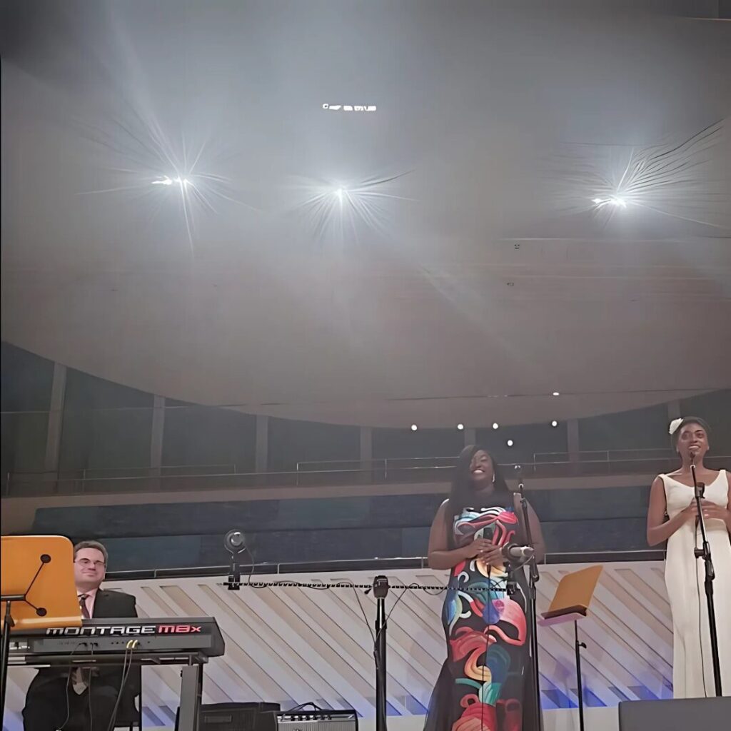 A musical performance on stage featuring a keyboard player on the left and two vocalists standing at microphones. The vocalist in the center wears a colorful patterned dress, and the vocalist on the right wears a white outfit with a headband. They are performing under bright stage lighting in a modern indoor venue with a curved ceiling and a minimalistic backdrop.