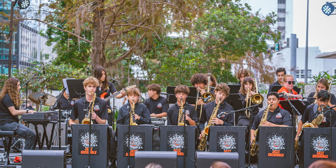 A color photograph of a large youth jazz ensemble performing outdoors on a stage in an urban setting. The group is composed of approximately fifteen students of varying ages, all wearing matching black T-shirts that say "JAZZ ENSEMBLE" on large speaker covers in front of the stage. The front row consists mainly of musicians playing saxophones, with others behind them playing instruments like the trombone, trumpet, electric guitar, drums, and keyboard. The musicians are focused on their sheet music and instruments. The background features lush green foliage and the tall, modern buildings of a city street.