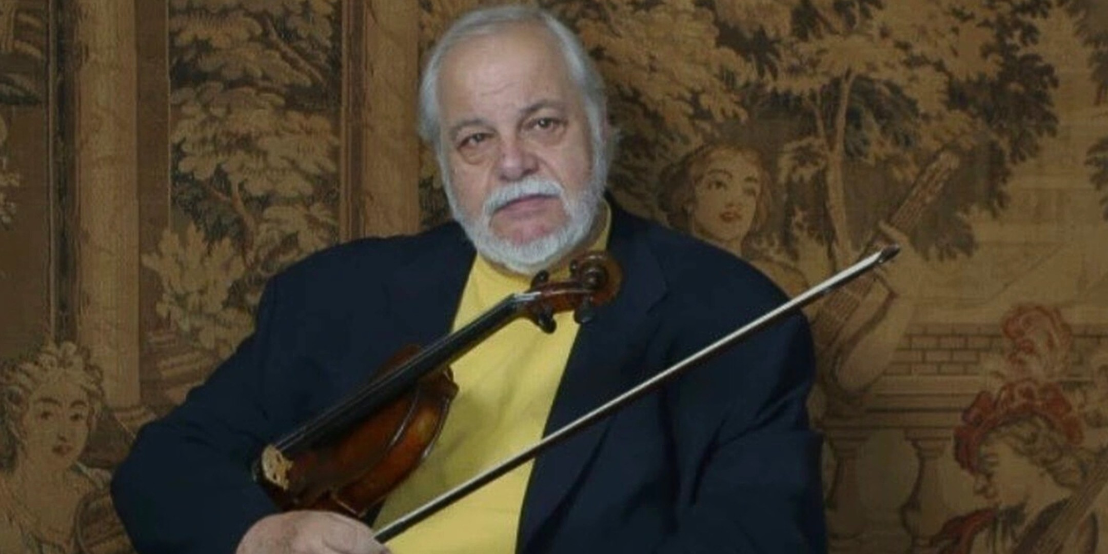 A color portrait of an older male musician, featuring a serious, direct expression. He has a white beard and gray hair and is wearing a dark navy blazer over a bright yellow shirt. He is holding a violin diagonally across his chest and has a violin bow in his right hand. The background is a rich, detailed, antique-style tapestry or woven wall hanging featuring scenes of figures and foliage in muted shades of brown, gold, and cream, suggesting a formal or historic setting.