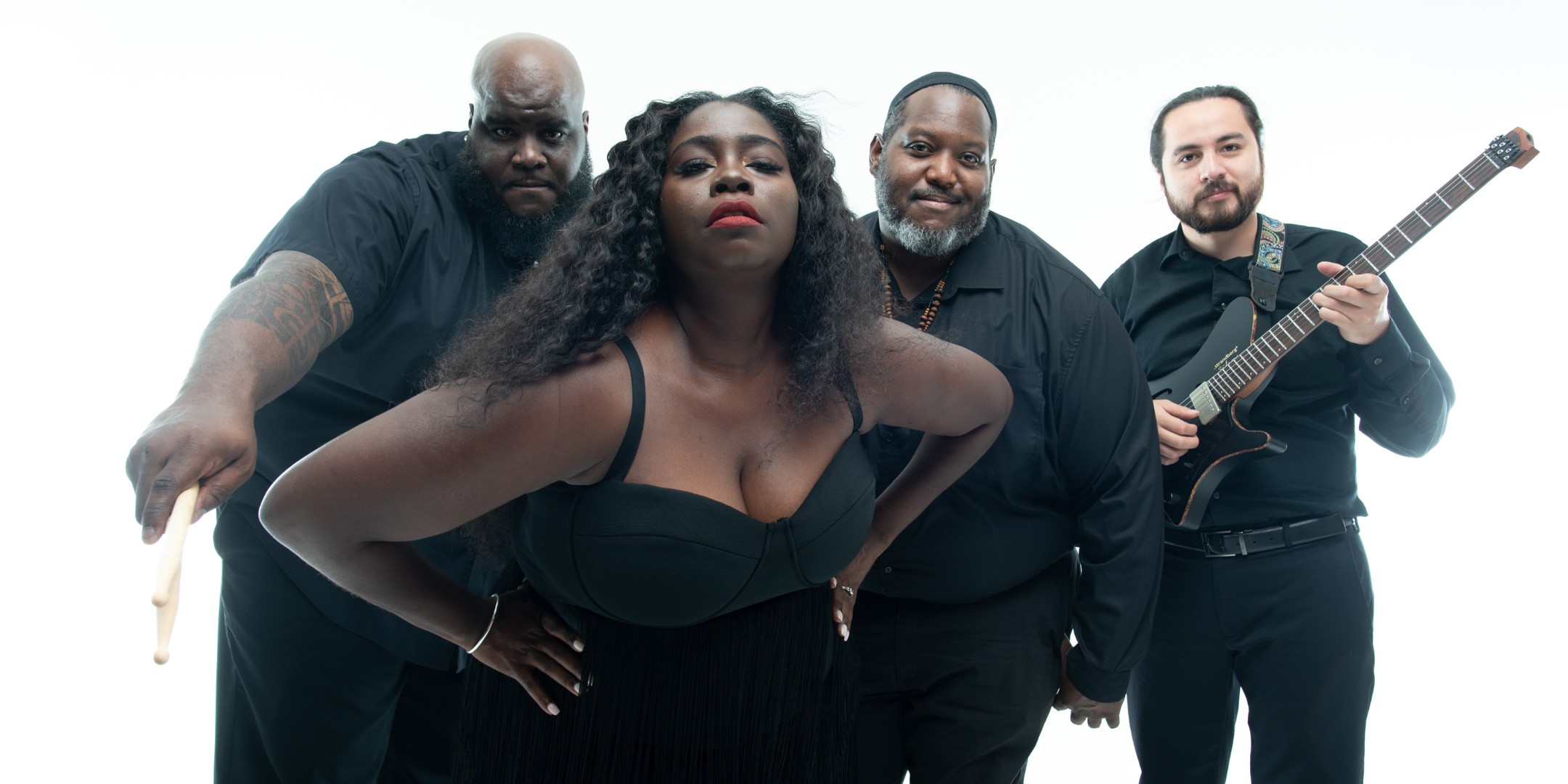 A dynamic, color studio portrait of a four-person band (The Harden Project), posing together against a pure white background. The group consists of one woman positioned prominently in the center foreground and three men slightly behind her. All four members are wearing black clothing. The female singer has long, dark, curly hair and wears a black bustier-style top with a fringed skirt, striking a confident pose with her hands on her hips and looking directly at the camera. The three male members include a drummer holding drumsticks, another member smiling slightly, and a fourth member holding a black electric guitar.
