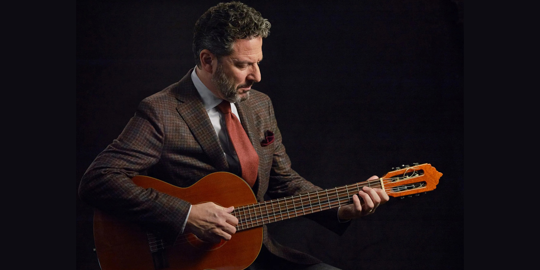 A studio portrait of a male jazz guitarist, focused intently on playing his acoustic guitar. He is seen from the chest up, seated against a dark, featureless background. The man has grey, curly hair and a beard, and wears an elegant, reddish-brown tweed sport coat and a matching burnt-orange tie. His eyes are directed downward at the neck of the guitar as his left hand fingers the strings and his right hand strums. The warm light highlights the texture of his jacket and the wooden body of the instrument.