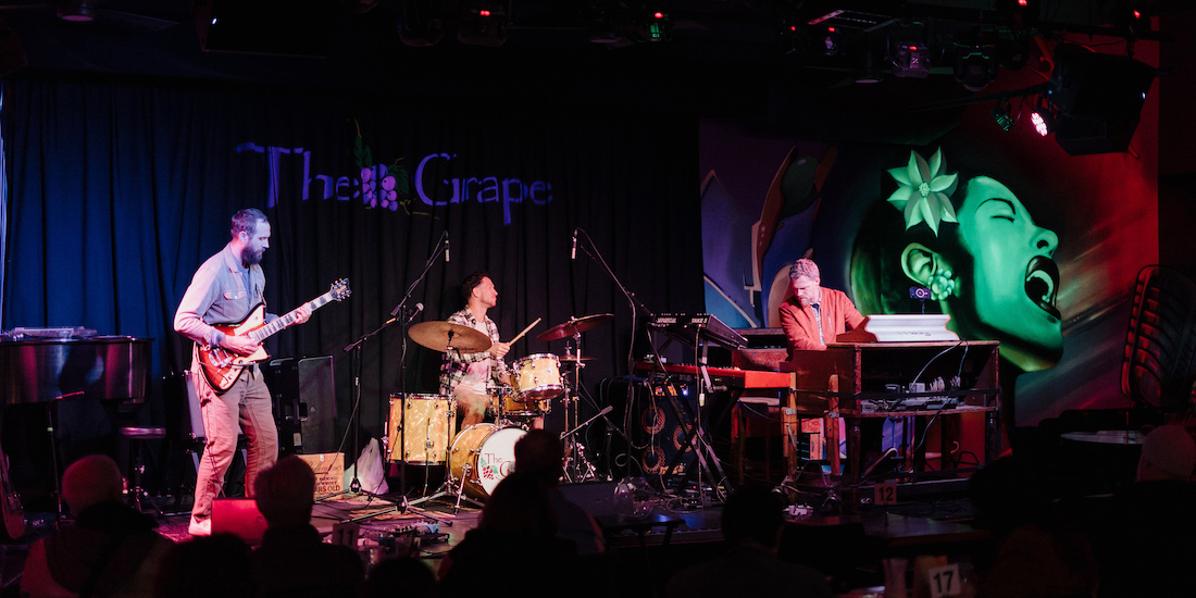 A color photograph capturing a live musical performance by a three-piece band (Lemon City Trio) on a dimly lit stage inside a music venue named "The Grape." The band members are playing their instruments: a guitarist stands on the left with an electric guitar, a drummer is seated in the center behind a drum kit, and a keyboardist is seated on the right playing a synthesizer. The stage is illuminated by colorful stage lights, casting blues and reds, which highlight a large mural on the right wall depicting a woman’s face with flowers in her hair. An audience is visible in the darkened foreground, watching the performance.