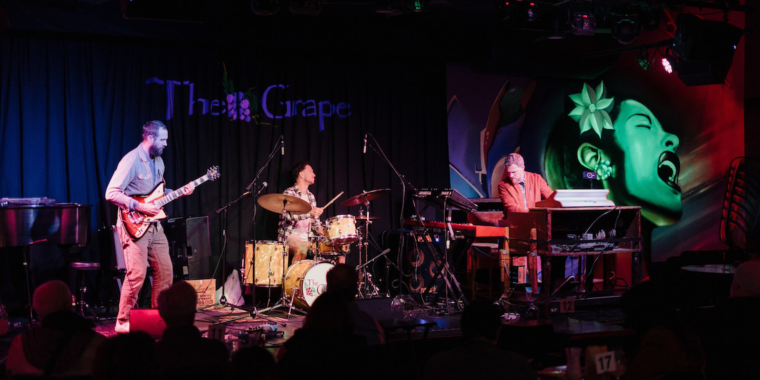 A color photograph capturing a live musical performance by a three-piece band (Lemon City Trio) on a dimly lit stage inside a music venue named "The Grape." The band members are playing their instruments: a guitarist stands on the left with an electric guitar, a drummer is seated in the center behind a drum kit, and a keyboardist is seated on the right playing a synthesizer. The stage is illuminated by colorful stage lights, casting blues and reds, which highlight a large mural on the right wall depicting a woman’s face with flowers in her hair. An audience is visible in the darkened foreground, watching the performance.