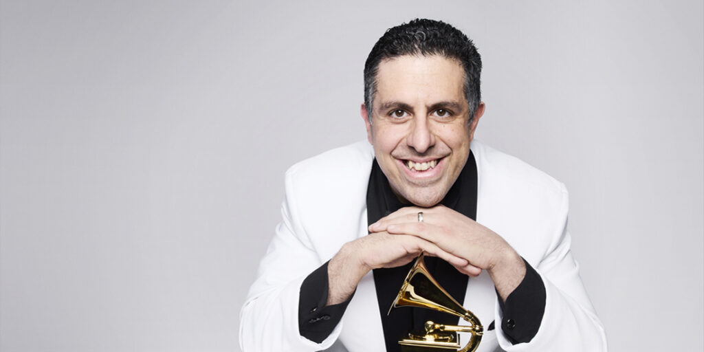 A brightly lit, color studio portrait of a male jazz historian or author, seated and looking directly at the camera with a wide, cheerful smile. He is wearing a contrasting outfit of a white jacket over a black shirt, and his dark hair is slicked back. His hands are clasped and resting on the bell of a small, brass Grammy Award trophy positioned directly in front of him. The background is a plain, solid light gray, causing the subject and the golden trophy to stand out clearly.