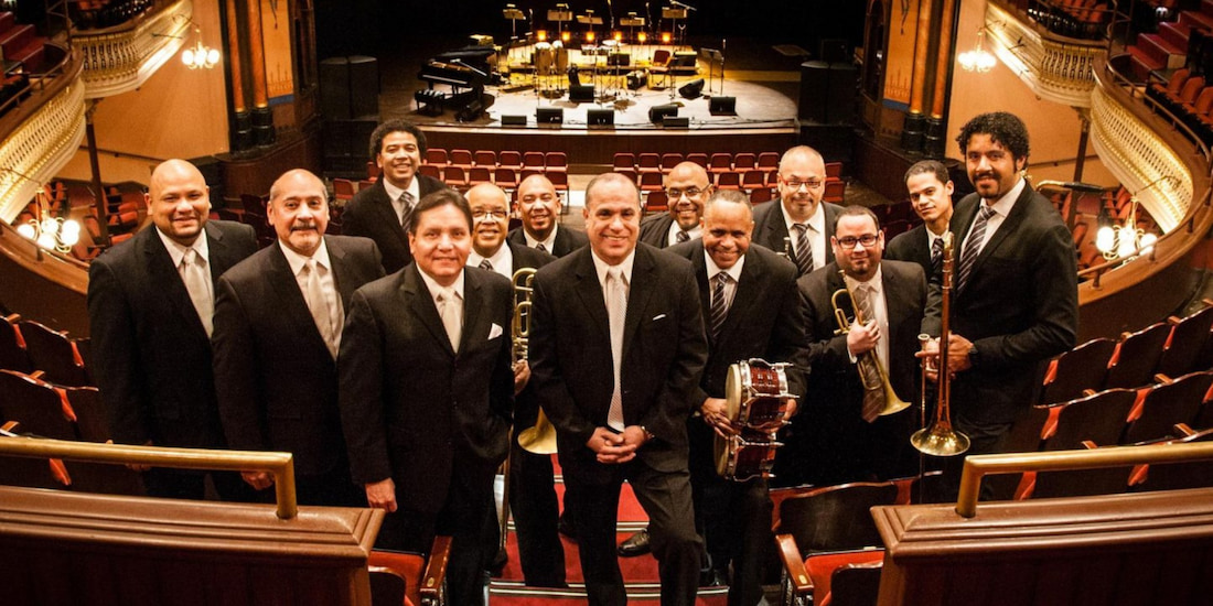 A promotional color photograph of a large, twelve-member Latin jazz or salsa orchestra posing formally inside a historic theater. All members are adult men dressed in matching black suits and light-colored ties. They are smiling and looking directly at the camera, conveying a feeling of professionalism and energy. Several members hold their instruments, including trumpets and a small hand drum. The band is positioned on the main floor, and the background shows the empty, tiered seating of the concert hall and the set-up stage equipment in the distance.