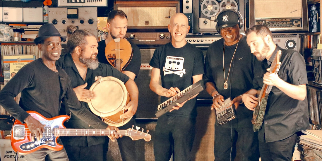 A color, wide-shot portrait of a six-member musical ensemble (The Spam Allstars) posing with their instruments in a cluttered studio or practice space. The group consists of six adult men, all dressed in dark casual clothing. The musicians are holding a variety of instruments: an electric bass guitar, a large hand drum (likely a tumbadora), a lute-like string instrument, a small electronic keyboard, and an electric guitar. The background is densely packed with vintage audio equipment, including reel-to-reel players, amplifiers, and various knobs and dials, giving the scene a retro or eclectic feel. The members appear relaxed and happy to be performing.