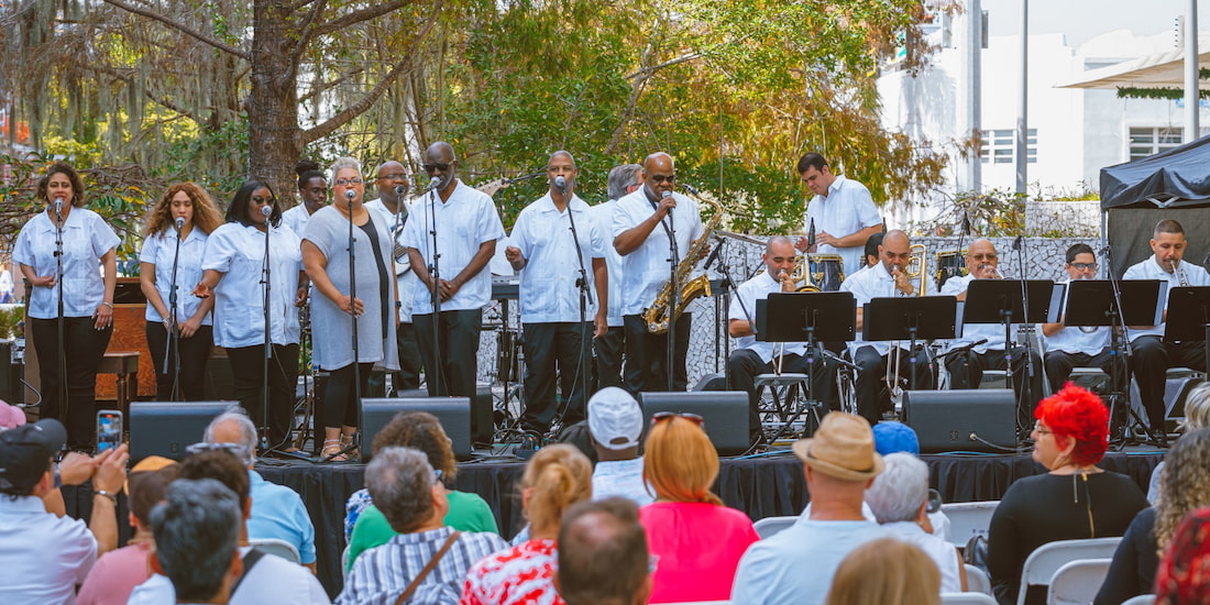 A wide-angle, color photograph showing a large gospel or jazz choir and band (Spirit of Goodwill) performing outdoors on a stage for a crowd. The performers are standing on a riser, mostly wearing matching white short-sleeved shirts and black pants. The group includes a line of singers holding microphones, and a brass section seated on the right side playing instruments like the trumpet and trombone, along with one standing member playing a saxophone. The audience is visible in the foreground, with many individuals wearing hats, seated and facing the stage. The setting is sunny and tropical, with large trees and Spanish moss visible behind the stage.