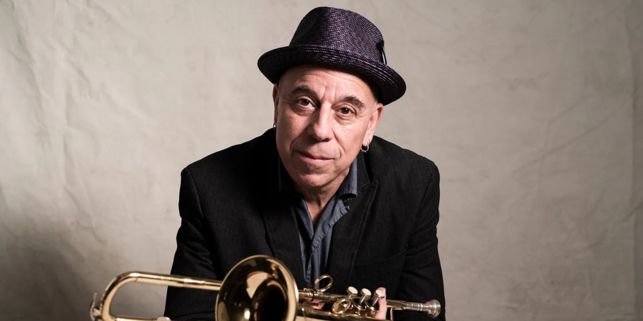 A close-up, studio color portrait of Steven Bernstein, a bald, middle-aged male musician, looking directly at the camera with a calm expression. He is wearing a dark, textured fedora or trilby hat, a navy jacket, and a light blue shirt. He holds a bright, brass slide trumpet or cornet horizontally beneath his chin, with the bell of the instrument pointing toward the lower left. The background is a soft, lightly wrinkled beige or off-white backdrop, providing gentle contrast to the darker colors of his attire.