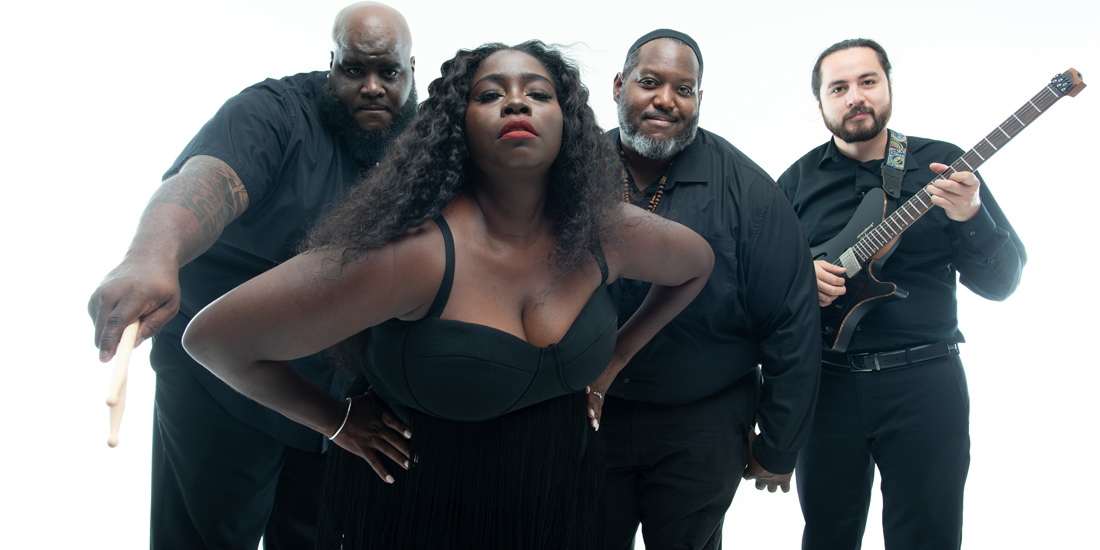 A dynamic, color studio portrait of a four-person band (The Harden Project), posing together against a pure white background. The group consists of one woman positioned prominently in the center foreground and three men slightly behind her. All four members are wearing black clothing. The female singer has long, dark, curly hair and wears a black bustier-style top with a fringed skirt, striking a confident pose with her hands on her hips and looking directly at the camera. The three male members include a drummer holding drumsticks, another member smiling slightly, and a fourth member holding a black electric guitar.
